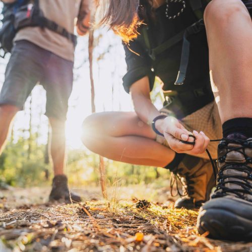 Close up of young female hiker tying shoelaces and getting ready for trekking in forest with man on background.